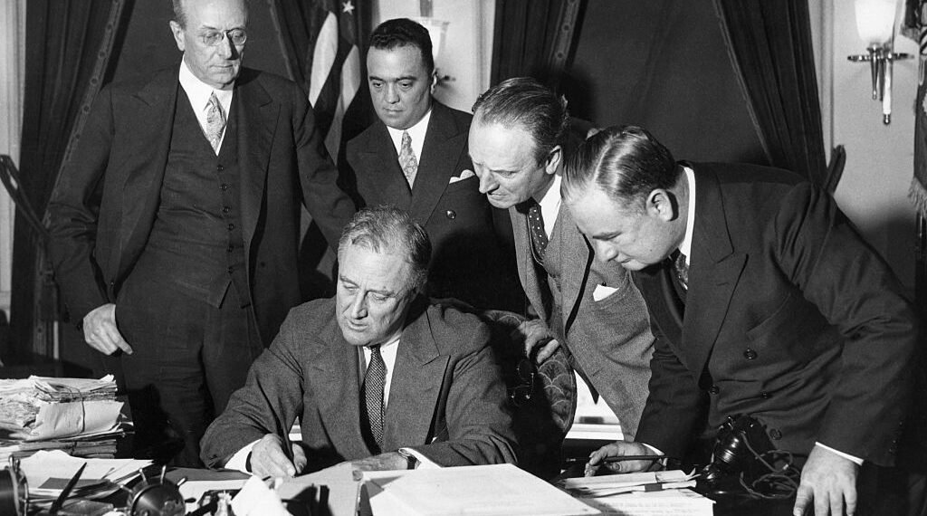 FBI Director J. Edgar Hoover stands behind President Franklin Delano Roosevelt and watches him sign a bill to enforce the fight against crime.